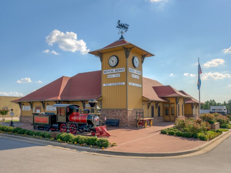 Exterior image of Dental Depot's South Tulsa dentist office with a clock tower, model train, flag pole, yellow exterior, and red roof.