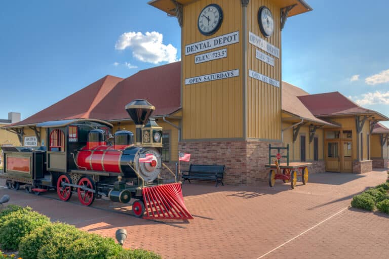 Exterior image of Dental Depot's South Tulsa dentist office with a clock tower, model train, yellow exterior, and red roof.