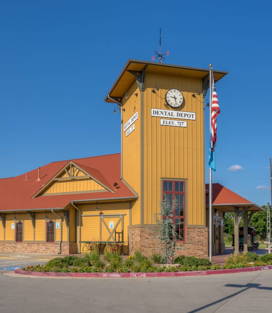 Exterior view of Dental Depot's Broken Arrow dentist office with a red roof, yellow walls, and a train station themed clock above the entrance.