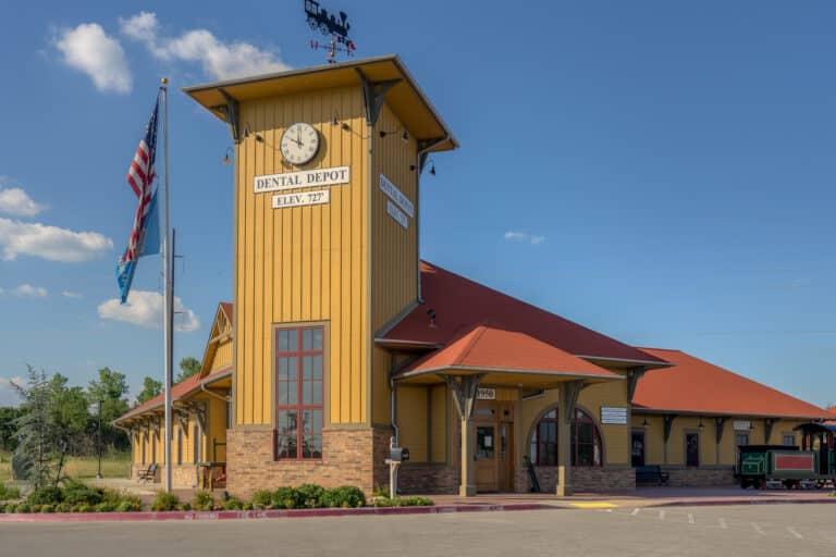 An exterior view of Dental Depot's Broken Arrow dentist office with yellow walls, a red roof, and a train station themed clock above the entrance.
