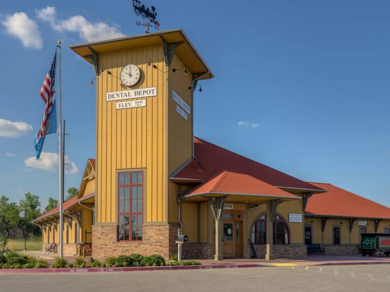 An exterior view of Dental Depot's Broken Arrow dentist office with yellow walls, a red roof, and a train station themed clock above the entrance.