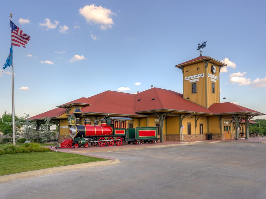 Exterior image of Dental Depot's Tulsa dentist office with a clock tower, model train, yellow exterior, and red roof.