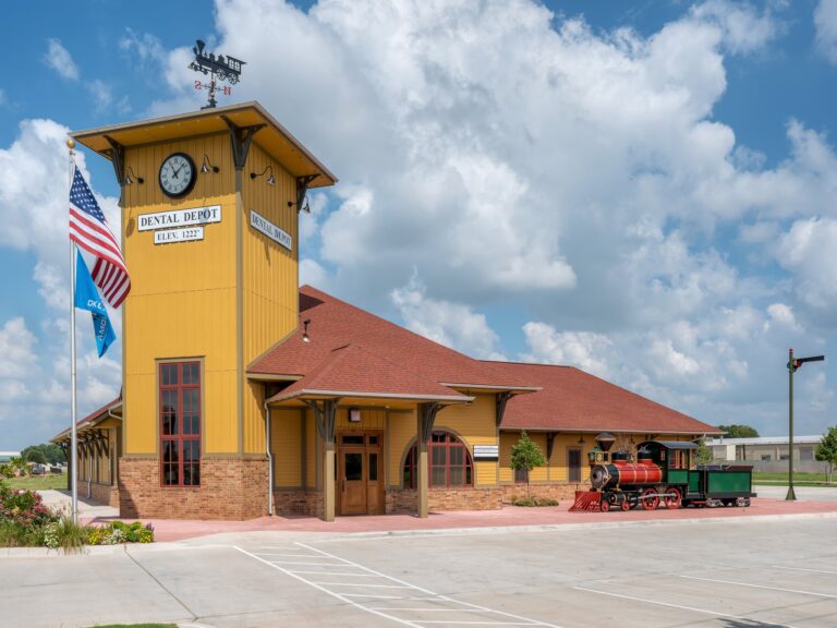 Exterior image of Dental Depot's North OKC dental clinic with a flagpole, clock tower, model train, and yellow exterior.