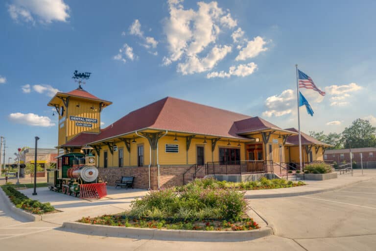 Exterior view of Dental Depot's Central Tulsa dental clinic with a model train, clock tower, and flagpole next to the yellow office.