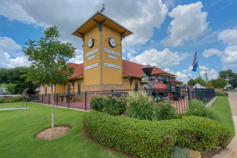 Exterior view of Dental Depot's Norman dental clinic with nice landscaping in the foreground and a building with a clock tower and a yellow exterior in the background.