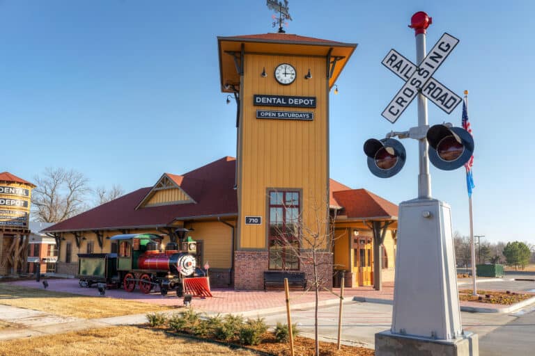 Exterior image of Dental Depot's Tulsa Hills dentist office with a clock tower, model train, train crossing sign, yellow exterior, and red roof.