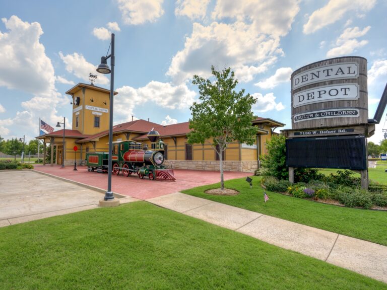 Exterior view of Dental Depot's Oklahoma City Dental Clinic on W. Hefner Rd with a model train, flagpole, water tower, and a yellow exterior.