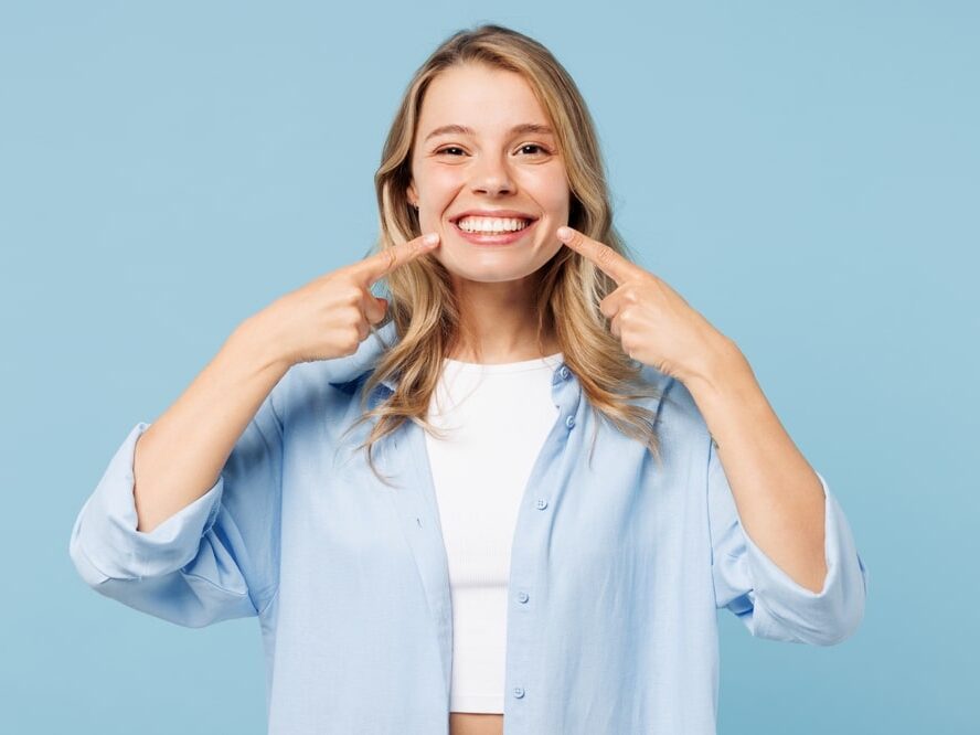 A blonde woman wearing a blue shirt points to her mouth while smiling broadly