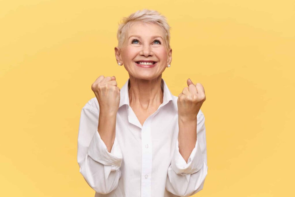 An older women smiles and pumps her fists on a yellow background.