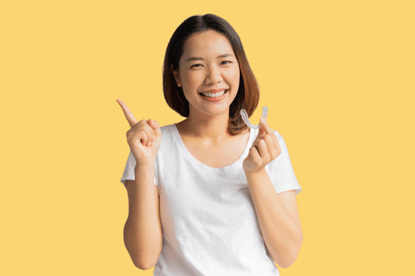 A young women smiles at the camera while holding an Invisalign Clear Aligner on a yellow background.