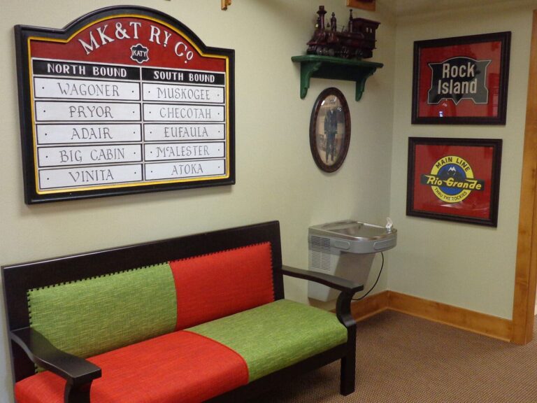 A second view of the lobby area of Dental Depot's dentist office in Broken Arrow with a train station themed destinations sign, pictures, a miniature train track, and seating.