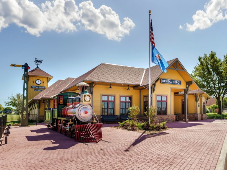 Exterior view of Dental Depot's Oklahoma City Dental Clinic on NW 63rd St. with a model train, flagpole, and a yellow exterior.