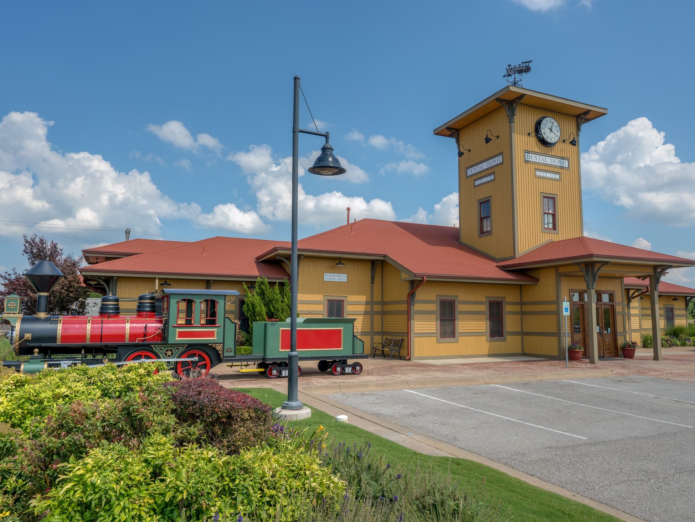 Exterior shot of the Edmond dental clinic with landscaping in the foreground, a model train, and a yellow exterior building with a clock tower.