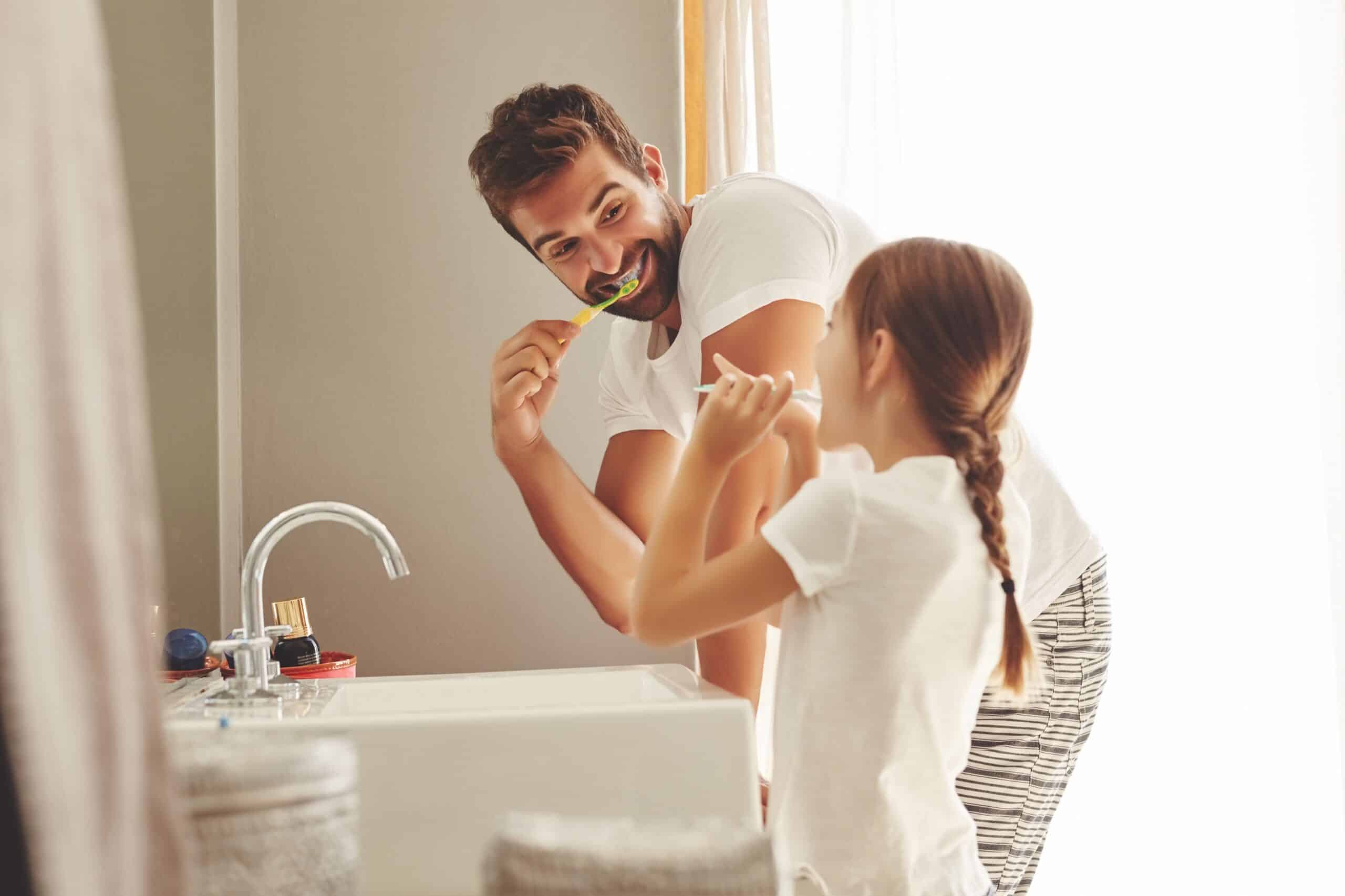 Father and daughter brush their teeth in bathroom of their home while smiling in the morning.