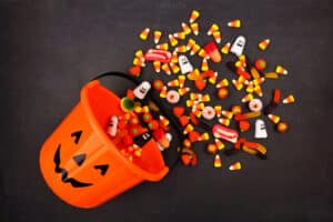 An orange bucket of Halloween Candy with a black, smiling jack-o-lantern face is spilling out candy on the floor.