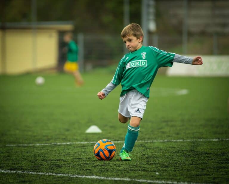 A young boy wearing green soccer gear is mid-kick on the field striking an orange soccer ball.
