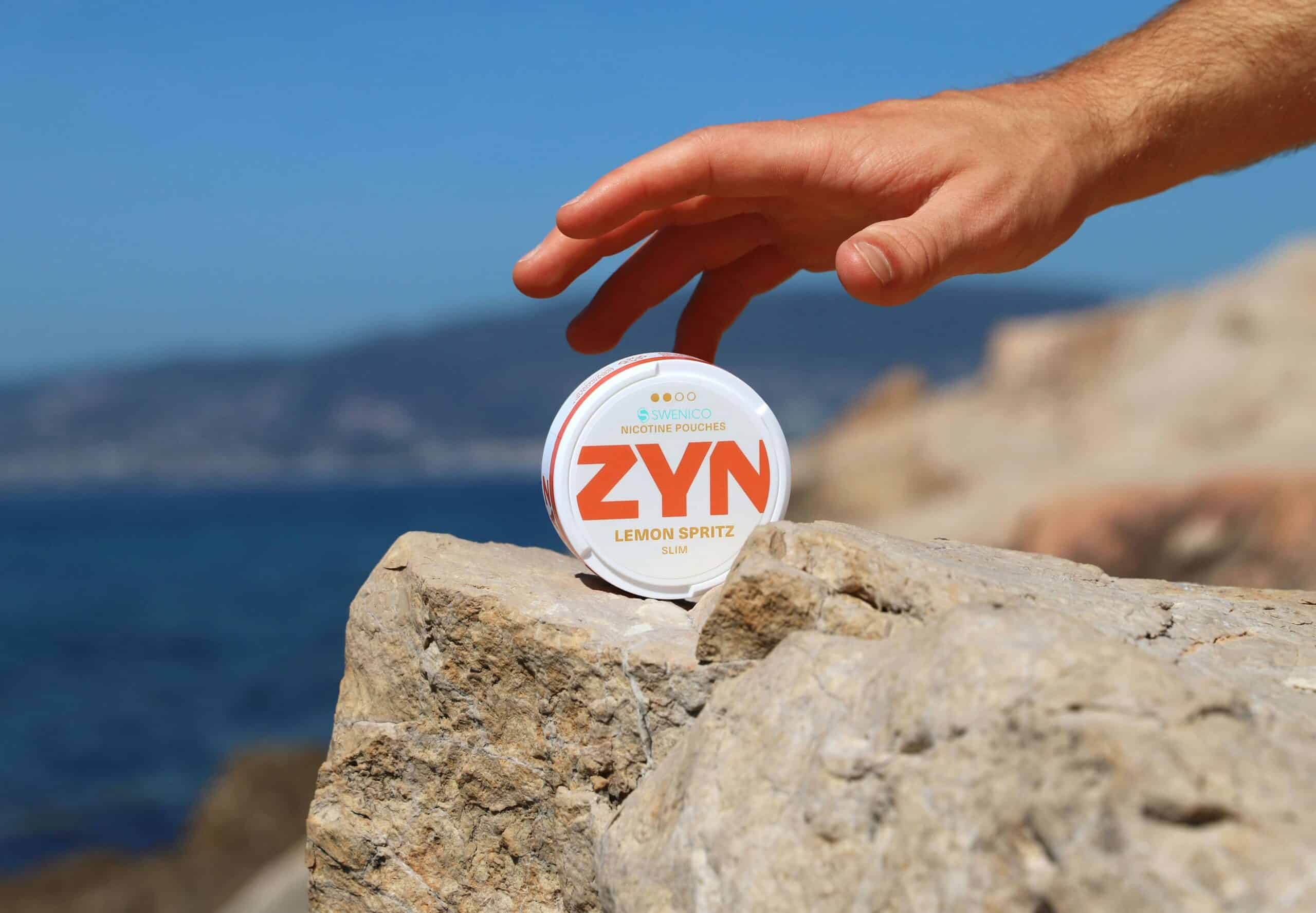 Hand reaching out for a ZYN tin of nicotine pouches, propped up on a stone next to the ocean.