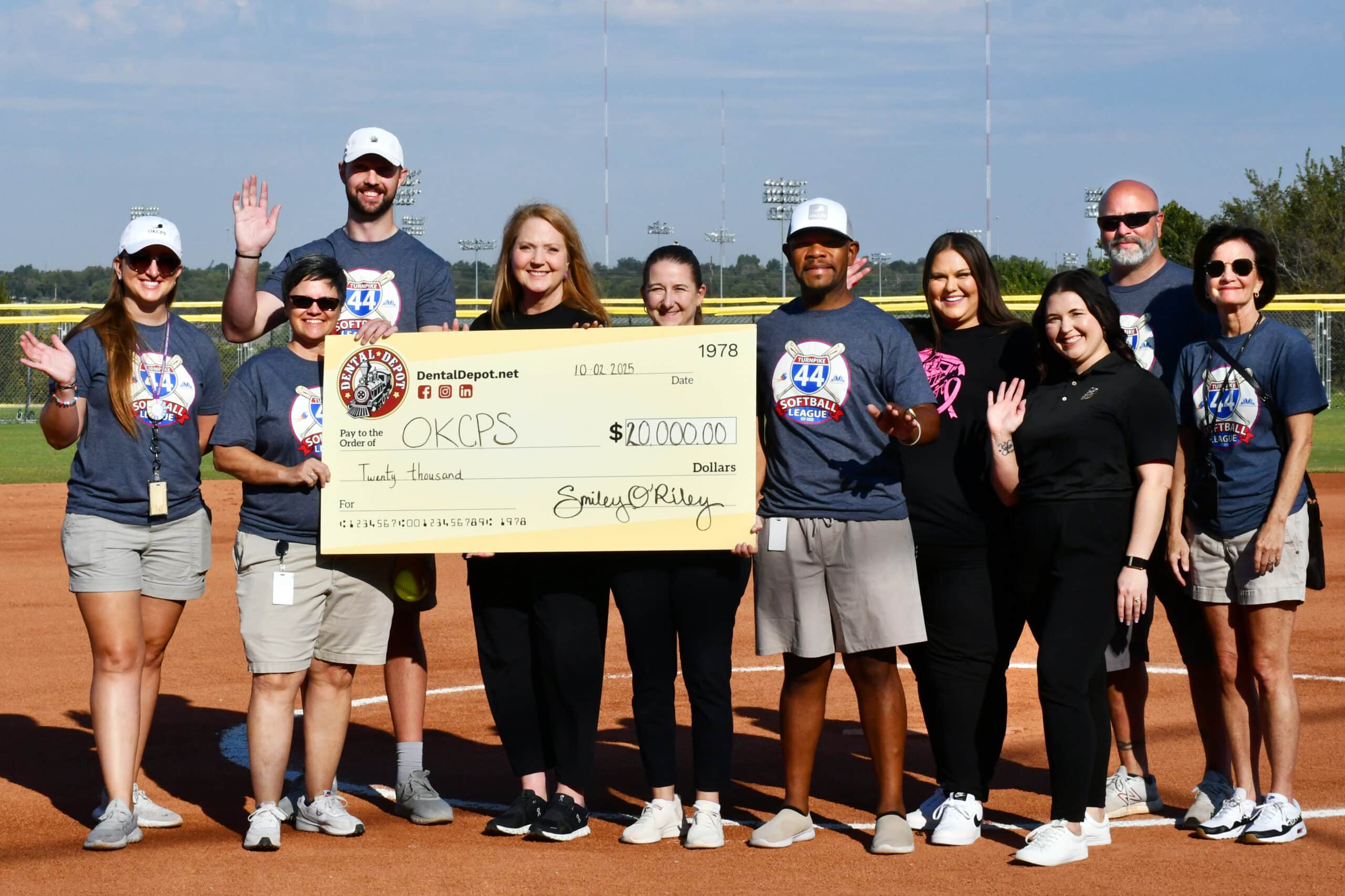 A representative of Dental Depot in OK gives a donation check to OKCPS outside on a baseball diamond.