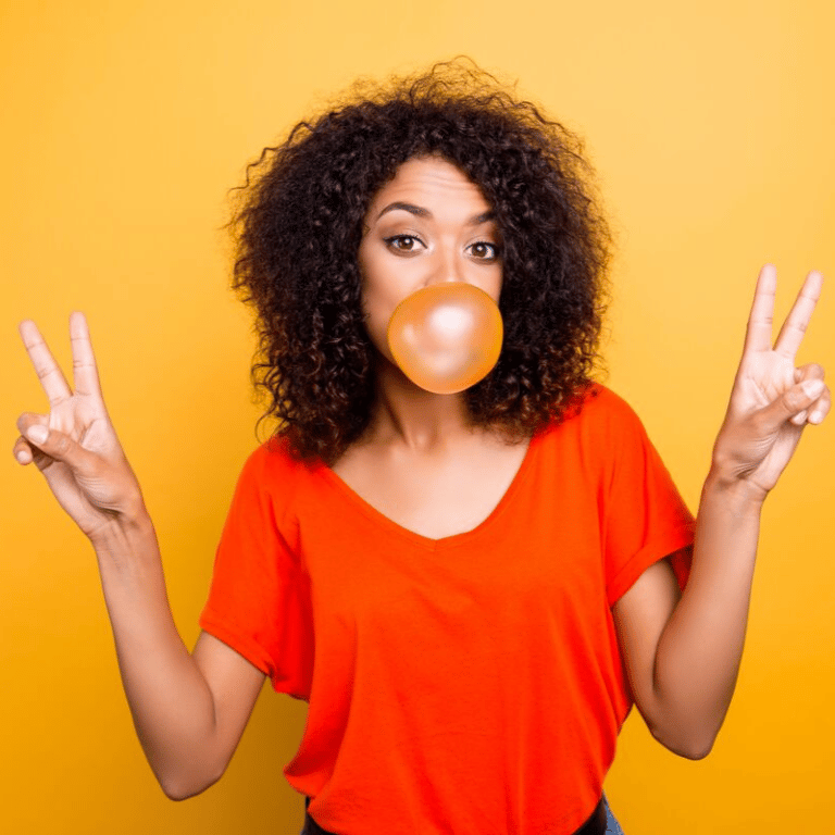 Image of a women with curly hair and a red shirt blowing a bubble with gum and giving the piece sign.