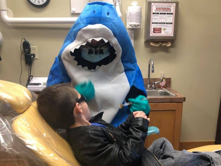 A dentist dressed in a shark costume works with a smiling child in the dental chair.