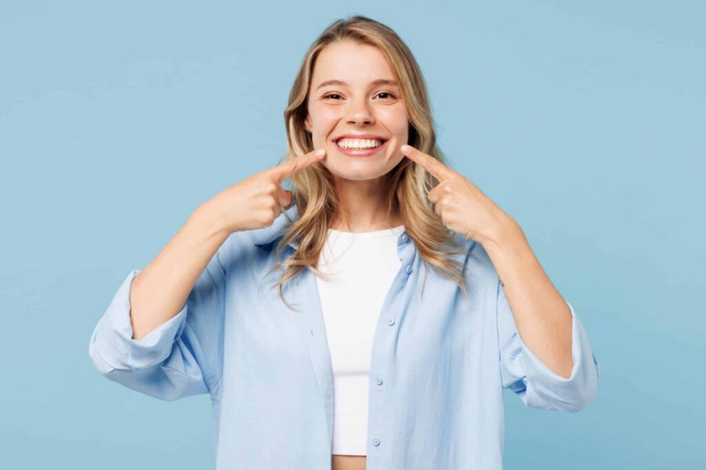 A young women smiles at the camera while pointing at her smile on a blue background.