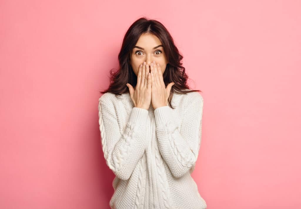 Brunette woman in a white sweatshirt covers her mouth to hide spots on her teeth in front of a pink backdrop.