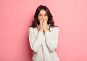 Brunette woman in a white sweatshirt covers her mouth to hide spots on her teeth in front of a pink backdrop.