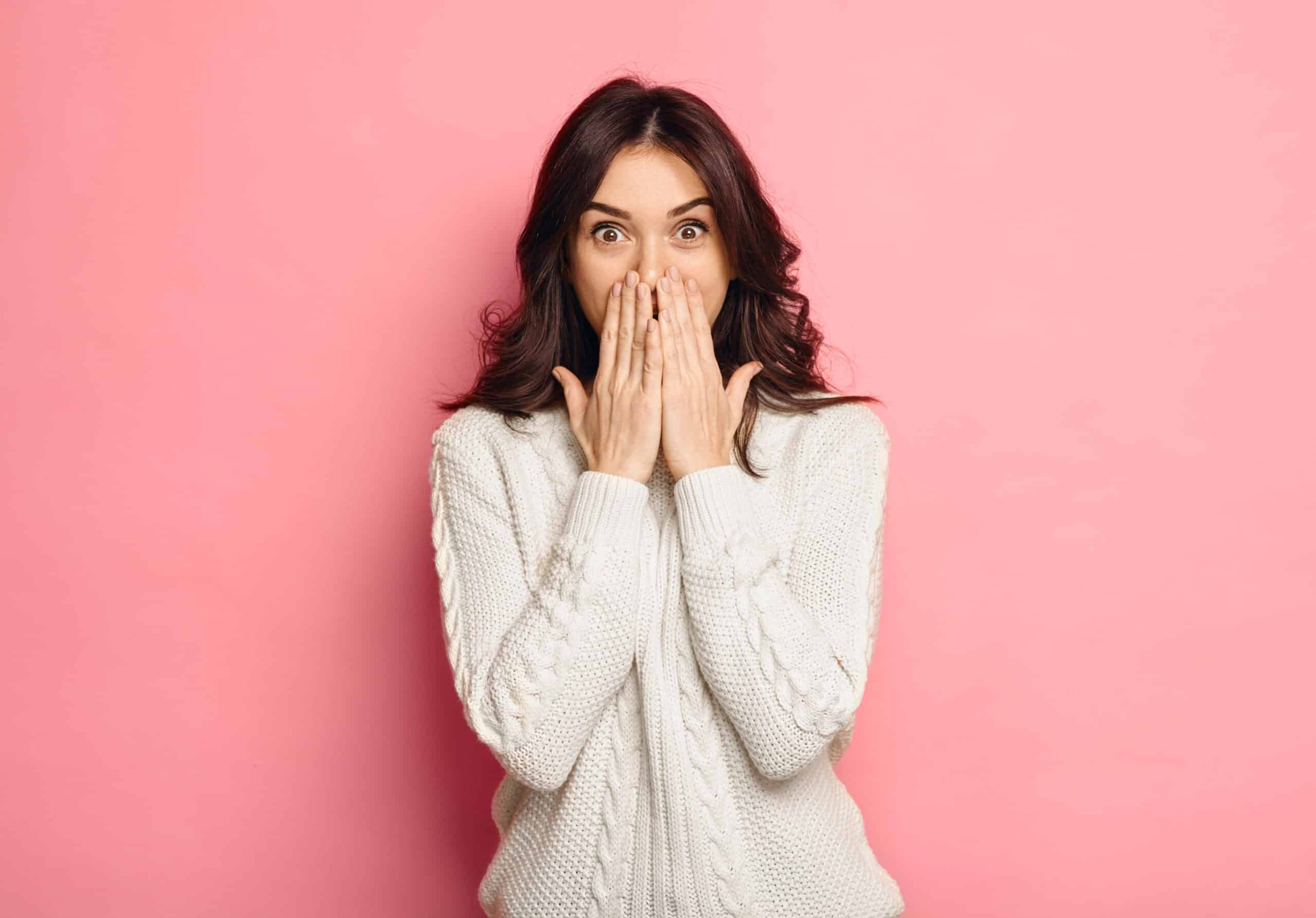 Brunette woman in a white sweatshirt covers her mouth to hide spots on her teeth in front of a pink backdrop.