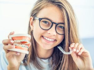A young woman holds up a model mouth with braces in one hand and Invisalign retainers in the other hand to compare them.