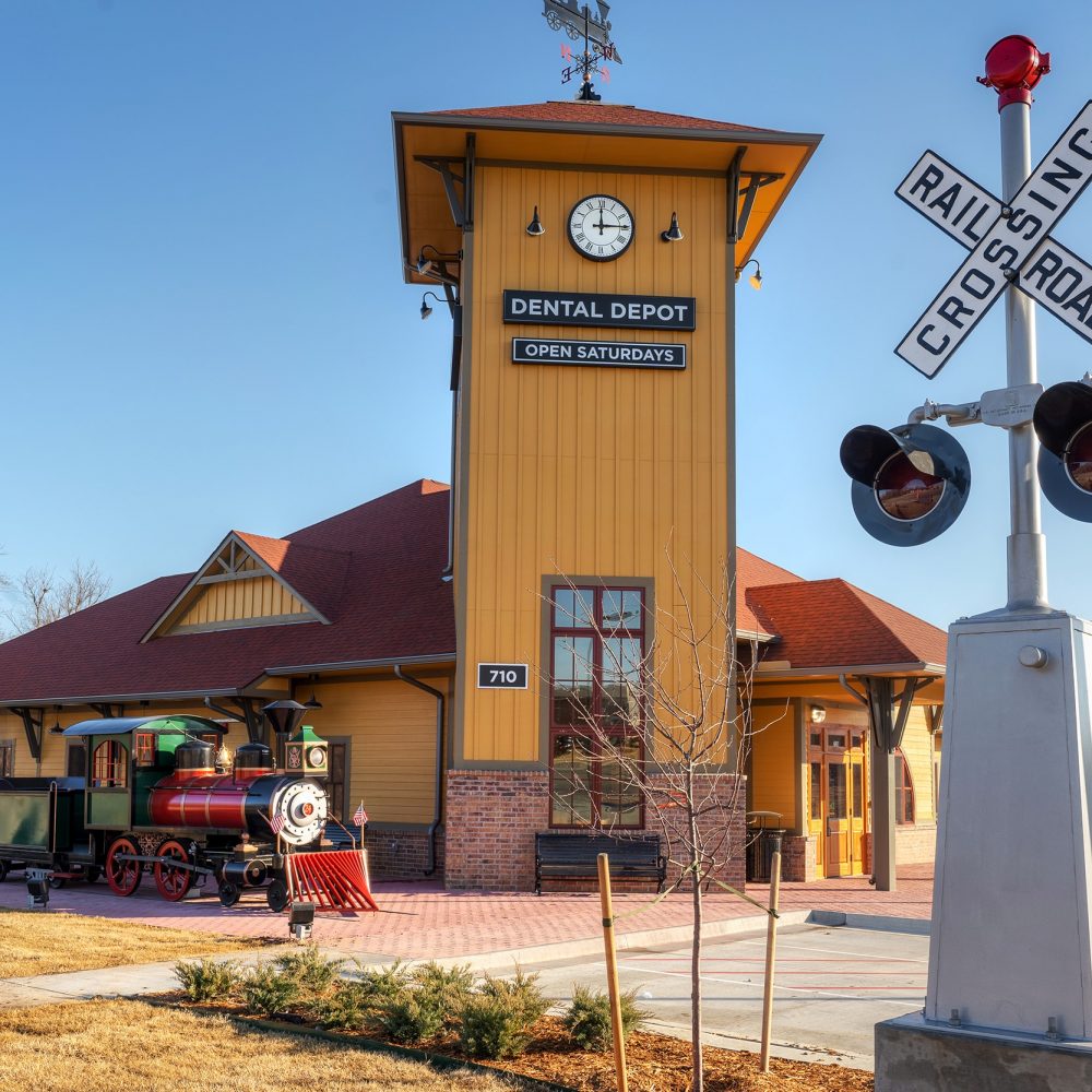 Exterior image of Dental Depot's Tulsa Hills dentist office with a clock tower, model train, train crossing sign, yellow exterior, and red roof.