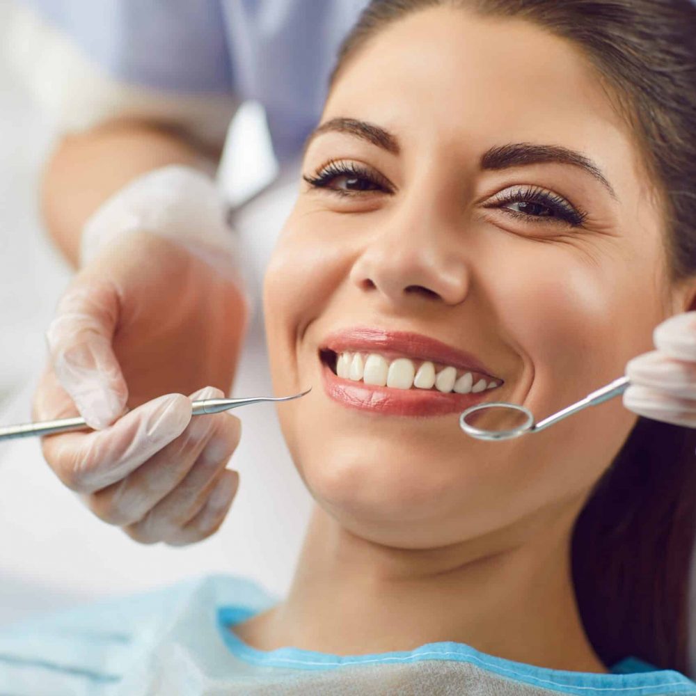 Brunette woman smiling while sitting in dental chair with dentist's hands holding teeth cleaning tools.