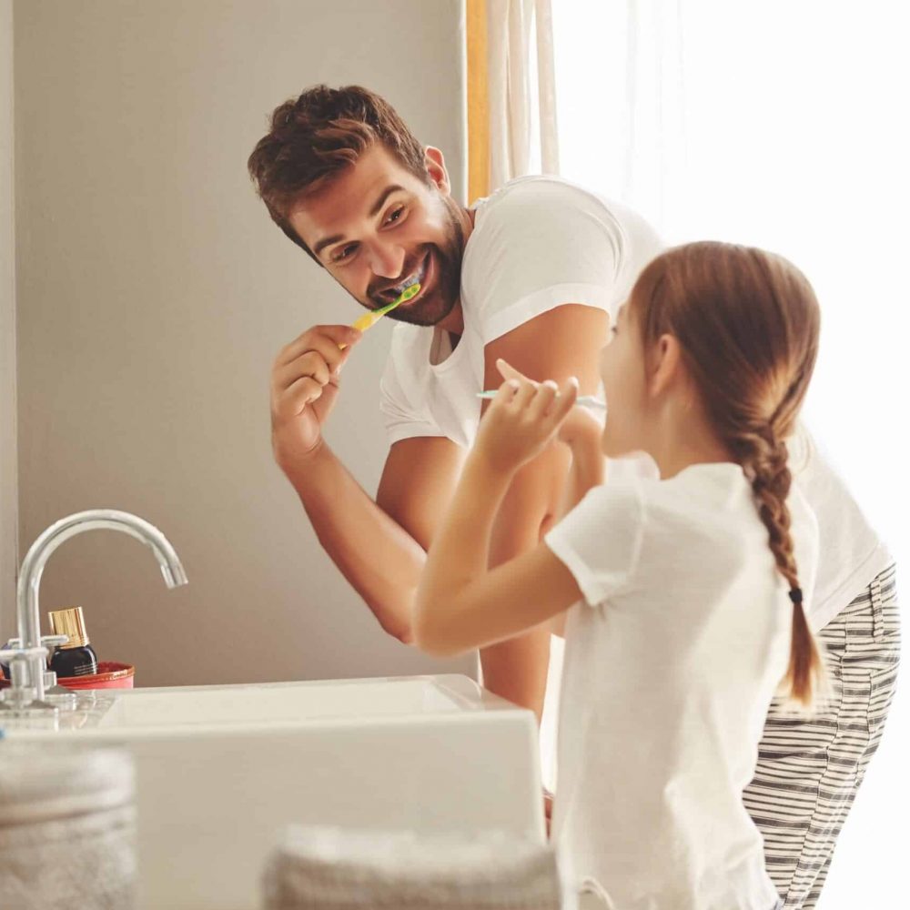 Father and daughter brush their teeth in bathroom of their home while smiling in the morning.