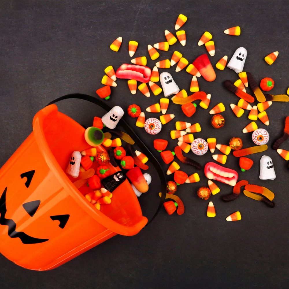 An orange bucket of Halloween Candy with a black, smiling jack-o-lantern face is spilling out candy on the floor.