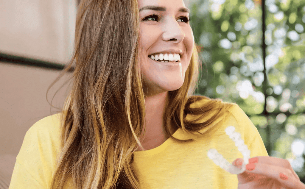 middle aged woman holding invisalign wearing yellow shirt and smiling.